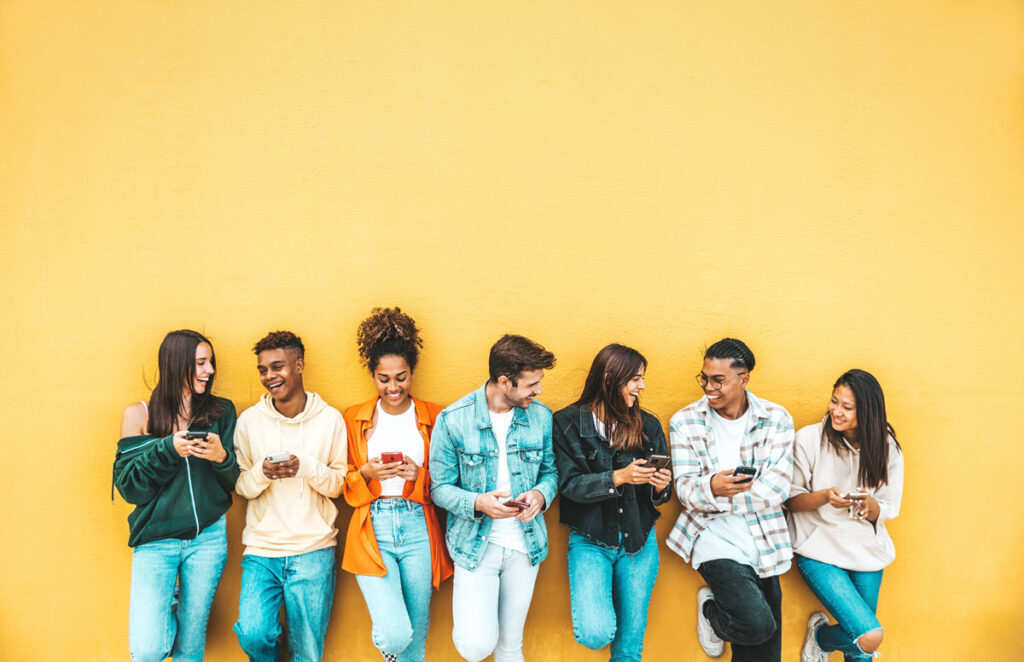 A group of people leaning against a yellow wall, holding and looking at their cell phones. 